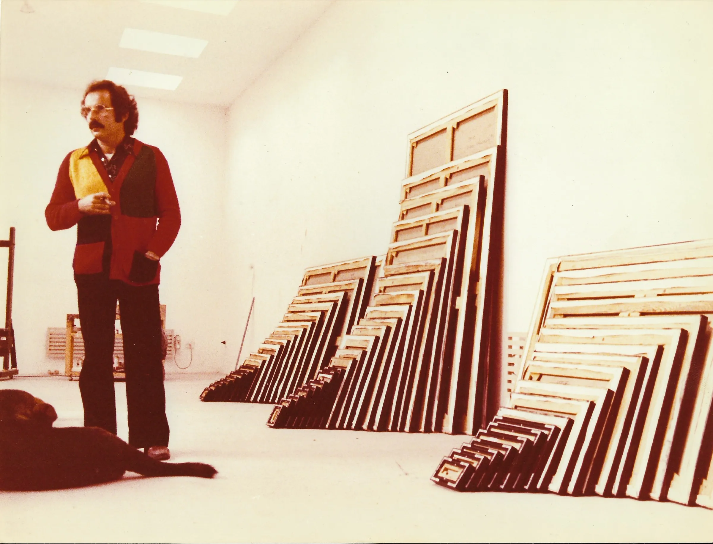 «Mides universals», 1979. Dret en una sala blanca amb lluernaris, armilla de punt vermella i groga; contra les parets, filades esglaonades de bastidors buits. A terra, un gos negre. Fotografia en color.