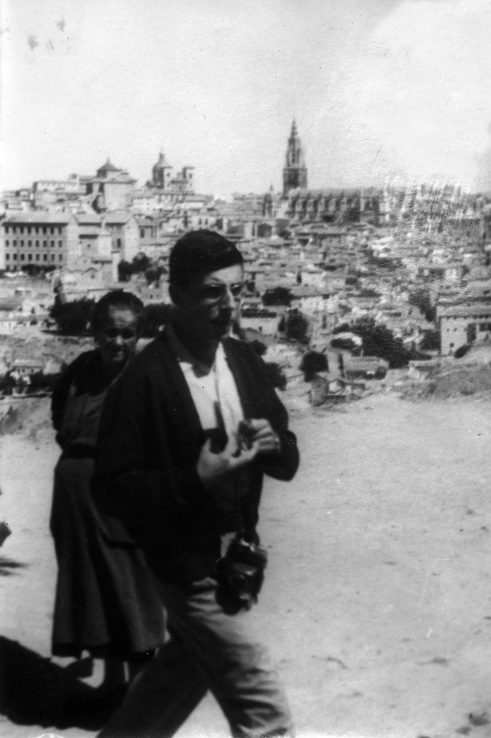 Caminant per un terreny sorrenc, amb una càmera penjada al coll; al fons, una ciutat en pendent i el campanar d'una catedral. Lloc sense identificar. Fotografia en blanc i negre.