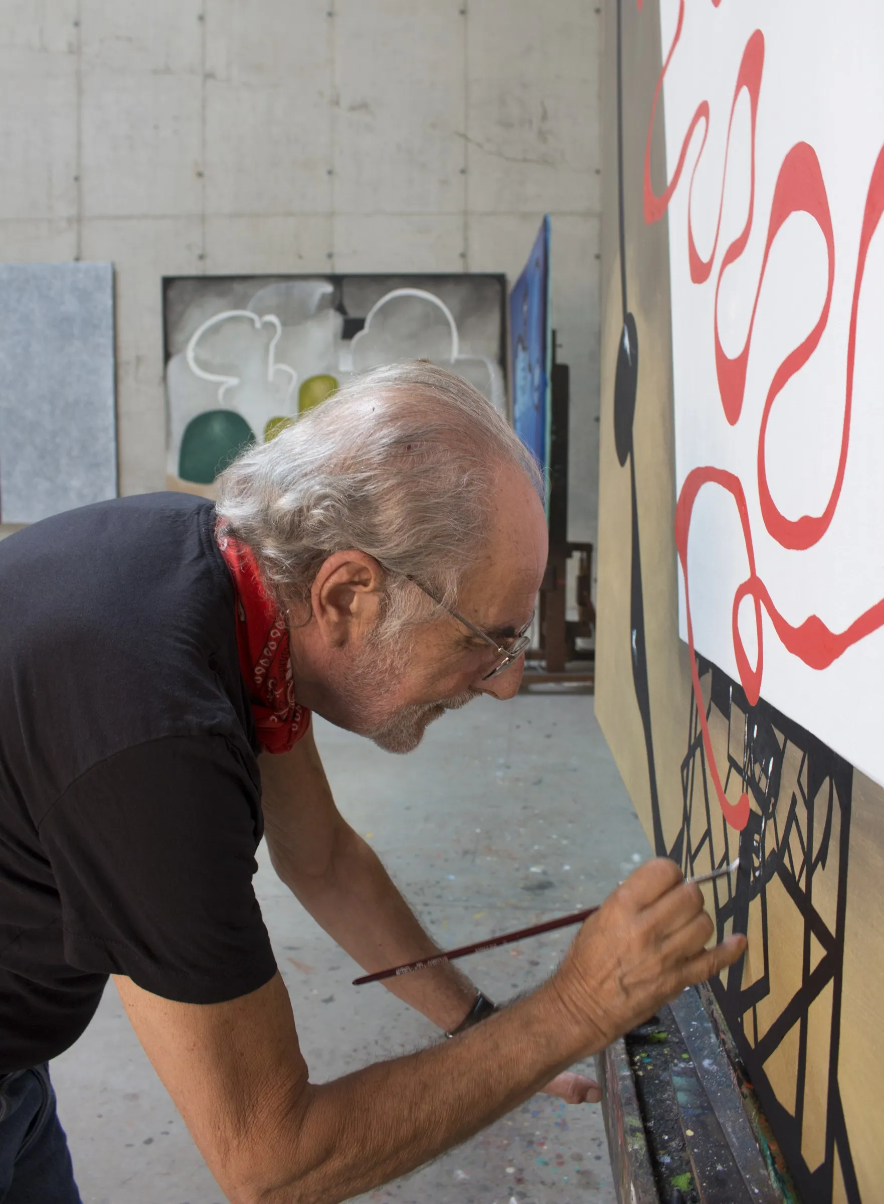 Eduard Arranz-Bravo pintant al taller de Vallvidrera, 2016: inclinat sobre la part baixa d'una tela gran —llaços vermells sobre fons blanc—, resseguint el traç negre amb un pinzell fi. Mocador vermell al coll; al darrere, la paret de formigó i obra recolzada.