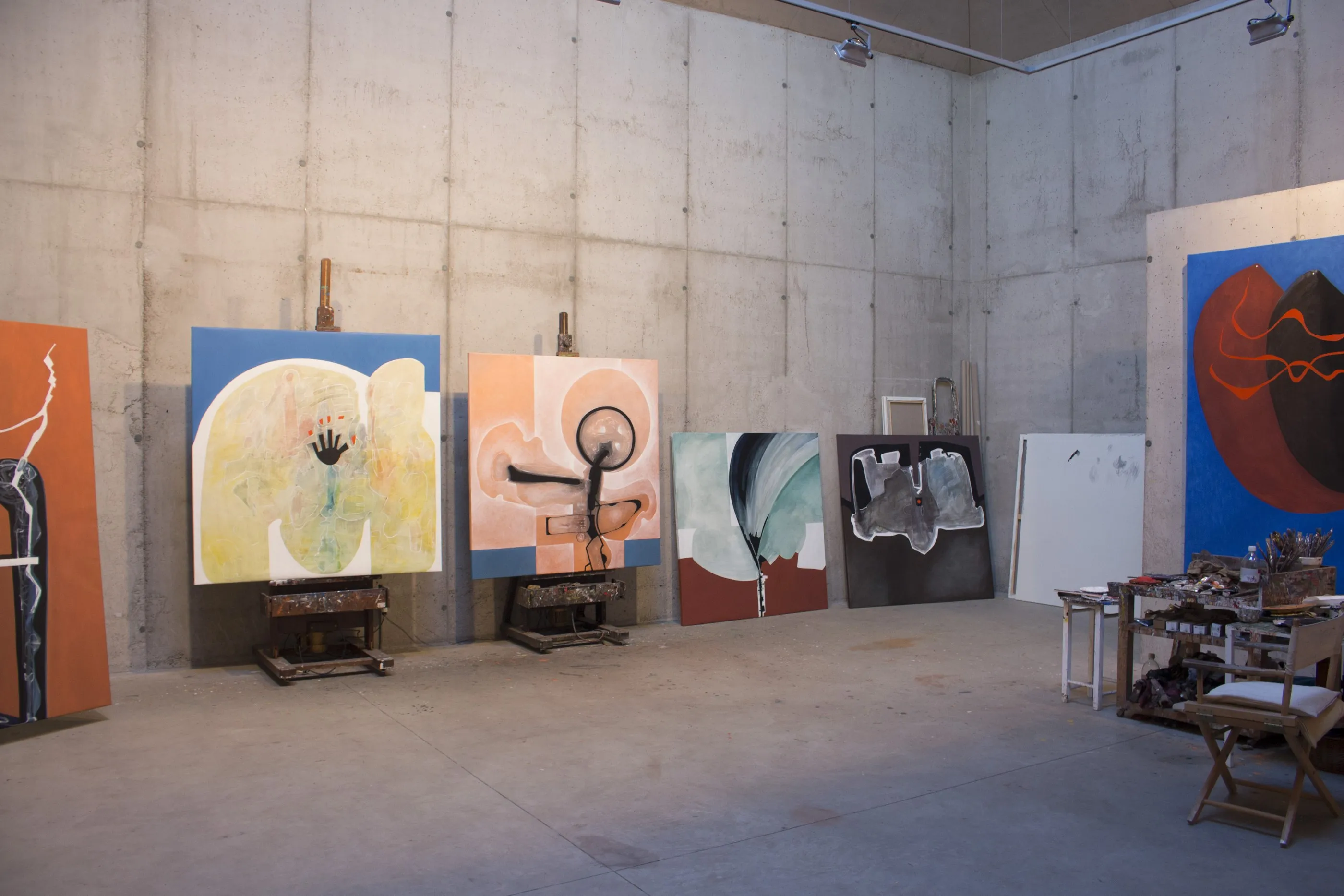 Racó de la sala de pintura amb dos cavallets de fusta antics que sostenen sengles teles: una de blava i groga amb una empremta de mà negra, i una de color salmó amb un cercle negre. A terra i contra la paret, més teles acabades i un plafó blanc buit.