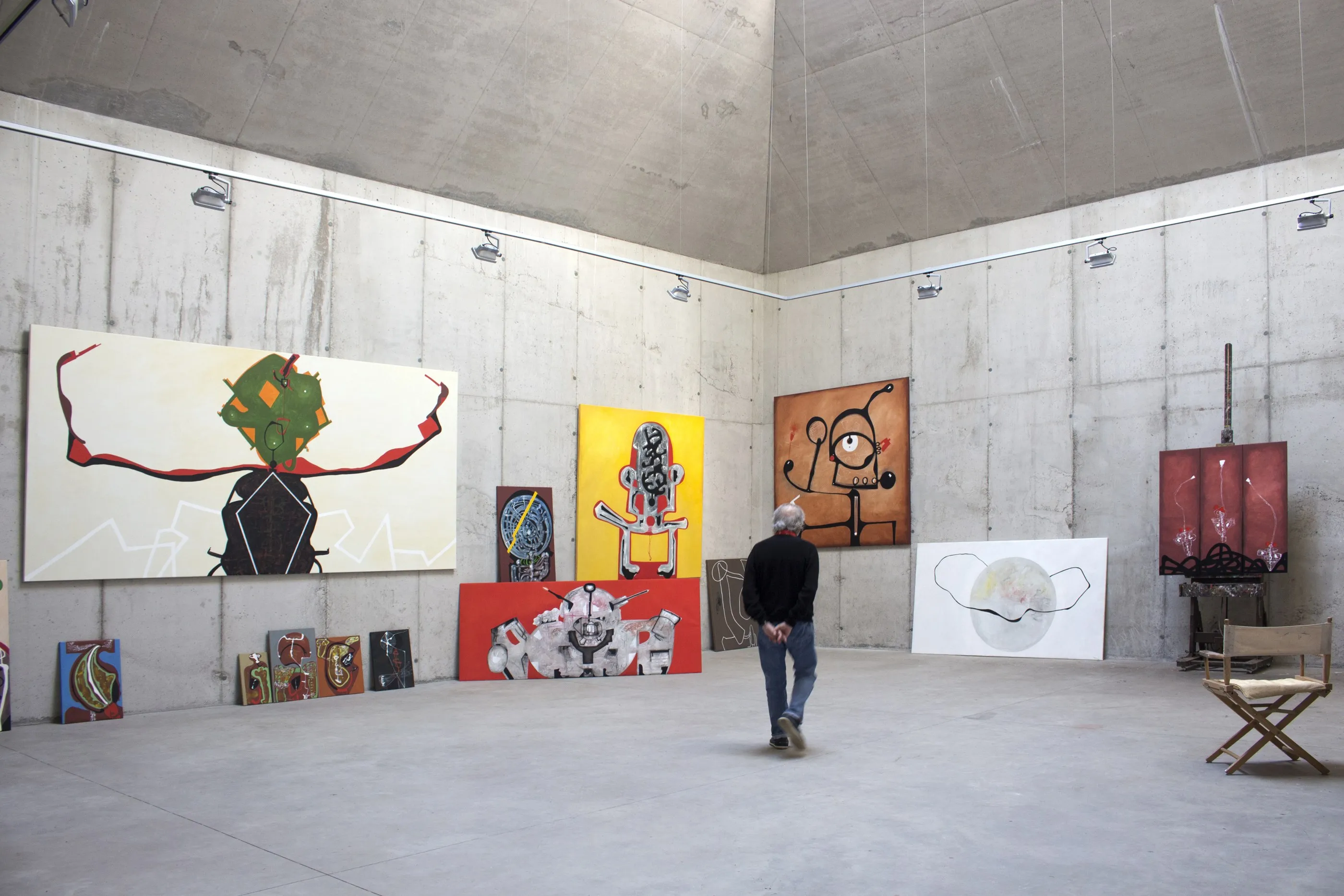La sala de pintura del taller de Vallvidrera. Nou obres de gran format a les parets de formigó vist, entre elles un políptic vermell fosc; al mig de la sala, Eduard Arranz-Bravo d'esquena, mirant-se l'obra amb les mans al darrere.