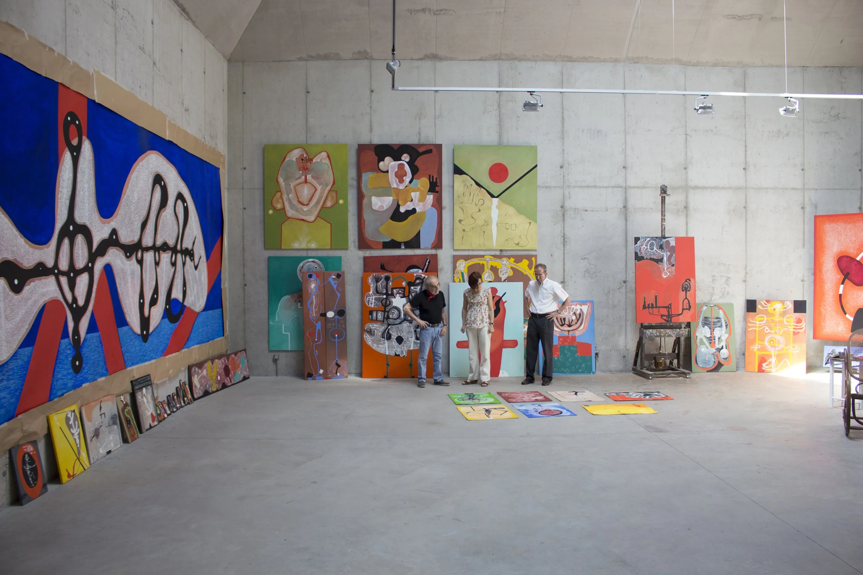 La sala de pintura durant una sessió de tria. A la paret del fons, sis teles quadrades penjades formant una graella. Tres persones dretes al centre miren avall, cap a una filera d'obres petites esteses a terra. A la paret esquerra, una tela molt gran blava i vermella subjectada amb paper.