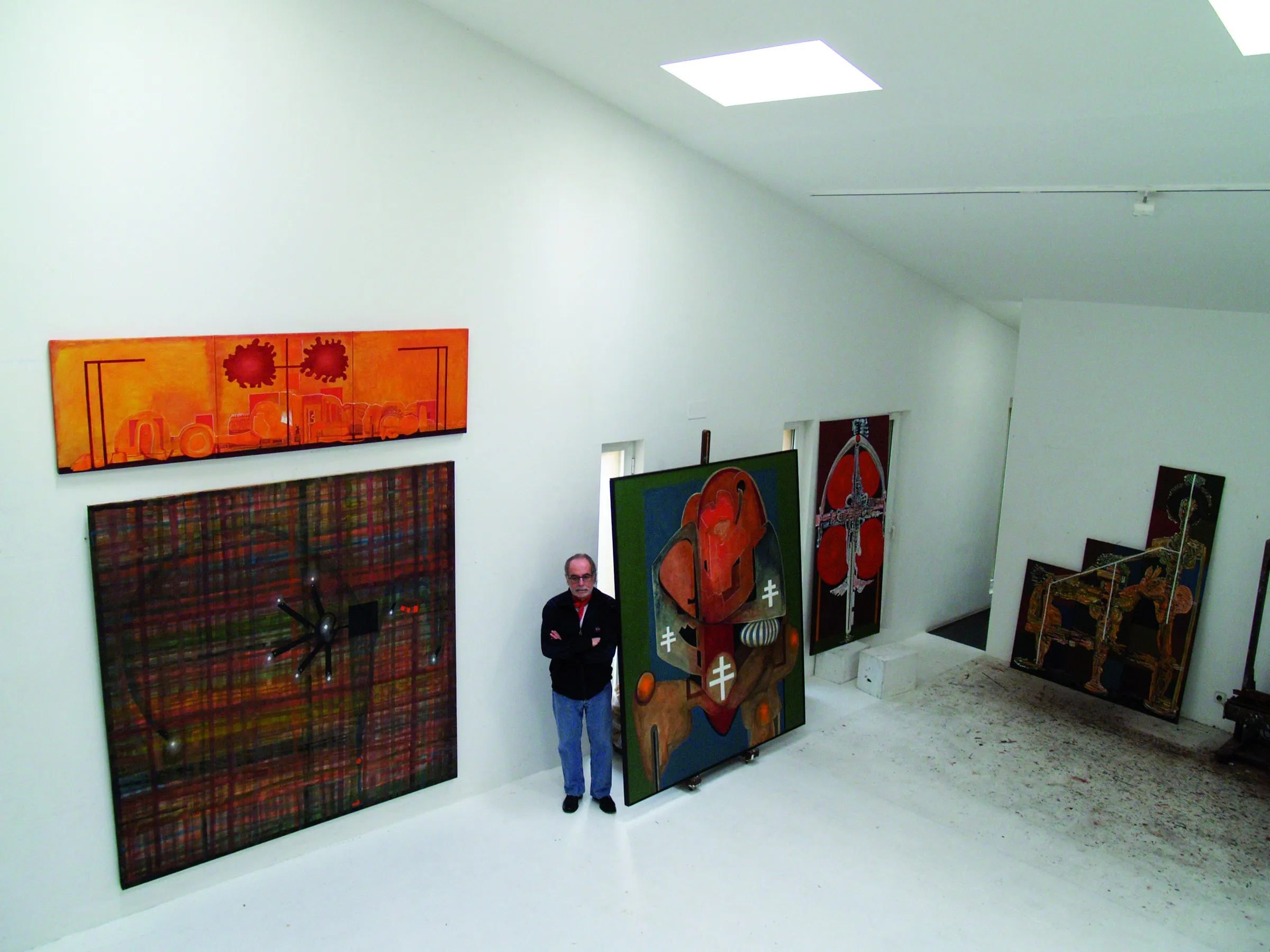 Sala de parets blanques amb un lluernari rectangular. Dret, amb els braços creuats, al costat d'una tela gran muntada damunt d'un carro; obra vermella i daurada recolzada a la dreta.
