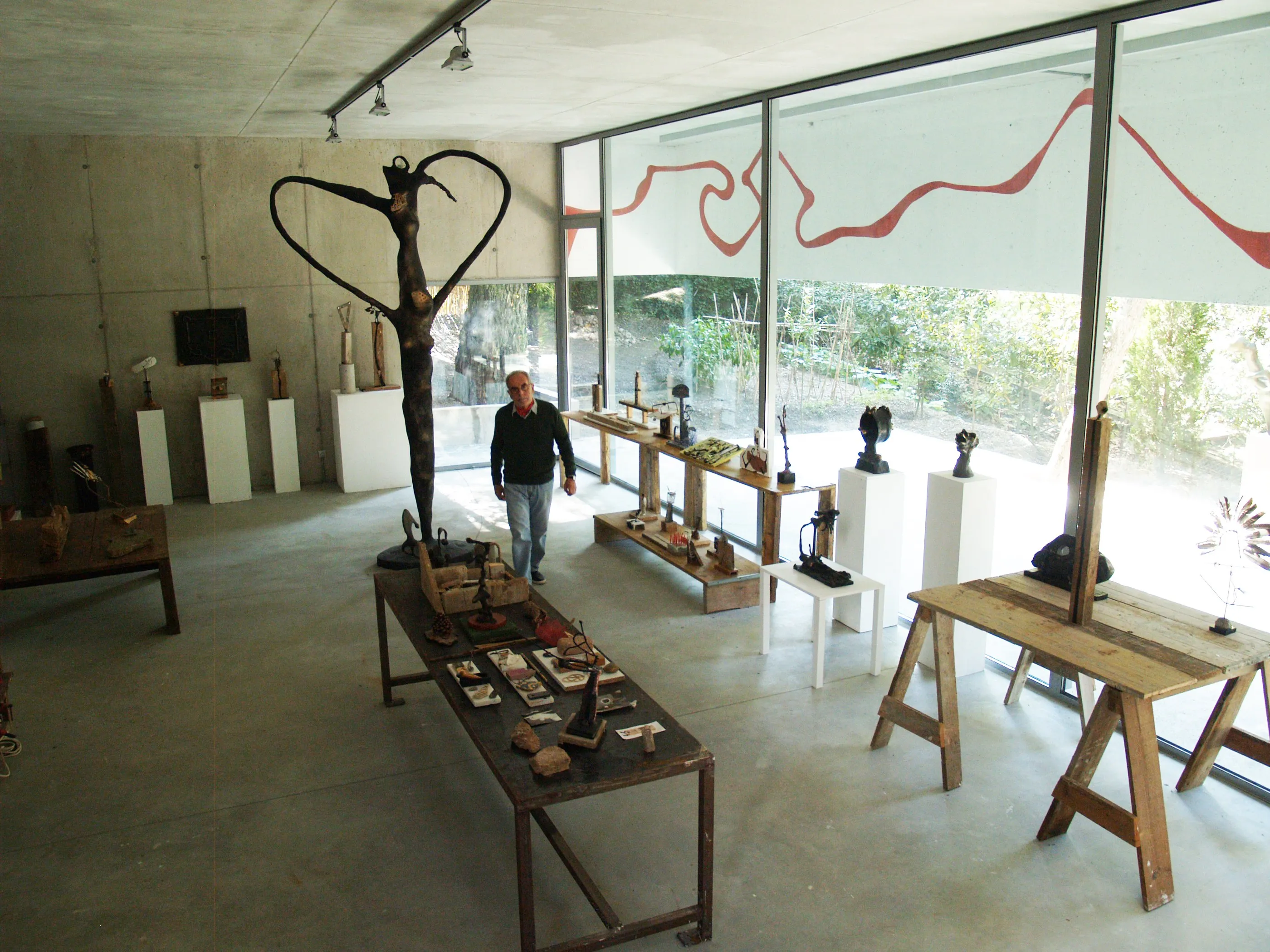El taller d'escultura vist des de dalt. A la dreta, una paret de vidre de tota l'alçada que dona al bosc, amb una línia vermella ondulant pintada damunt dels vidres i que travessa totes les fulles. Al centre, una escultura alta de bronze fosc amb els braços corbats formant un llaç. Taules de cavallets amb bronzes petits, maquetes i eines.